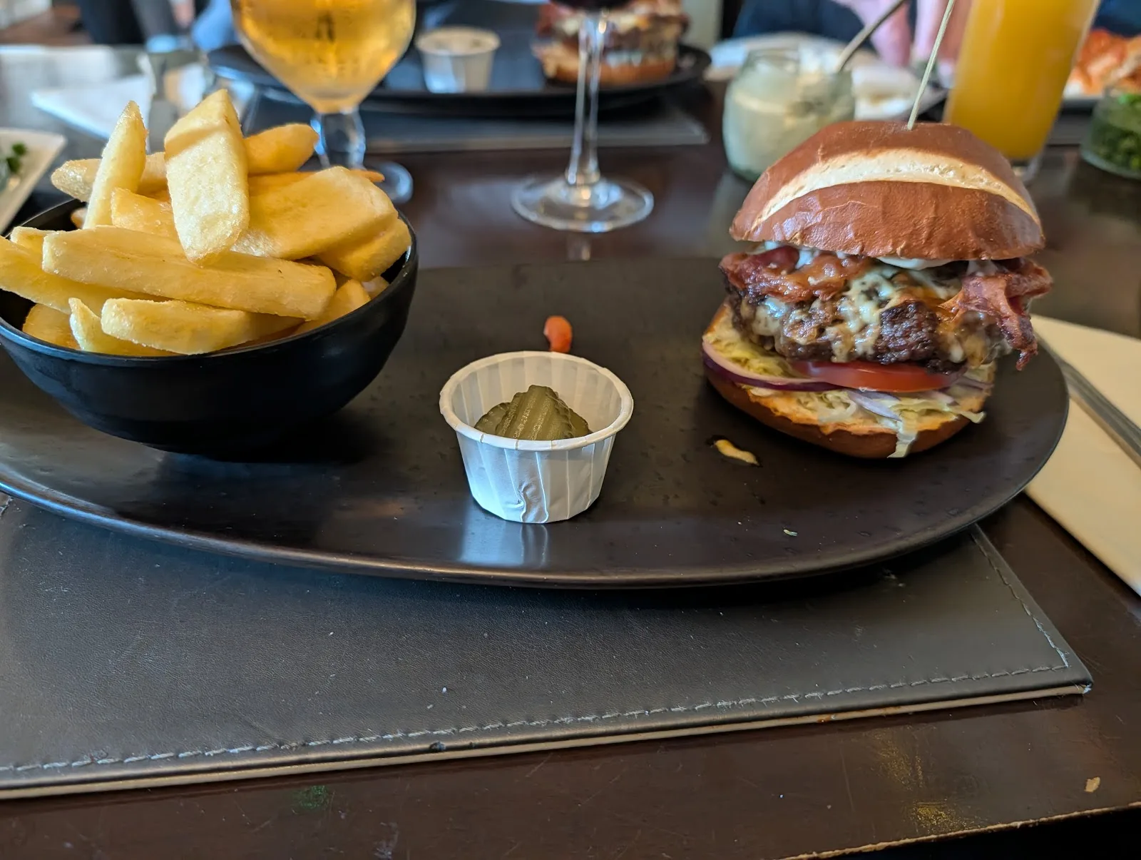 A bacon and cheese burger in a glazed bun with a bowl of chunky chips and a gherkin at The Olive Leaf