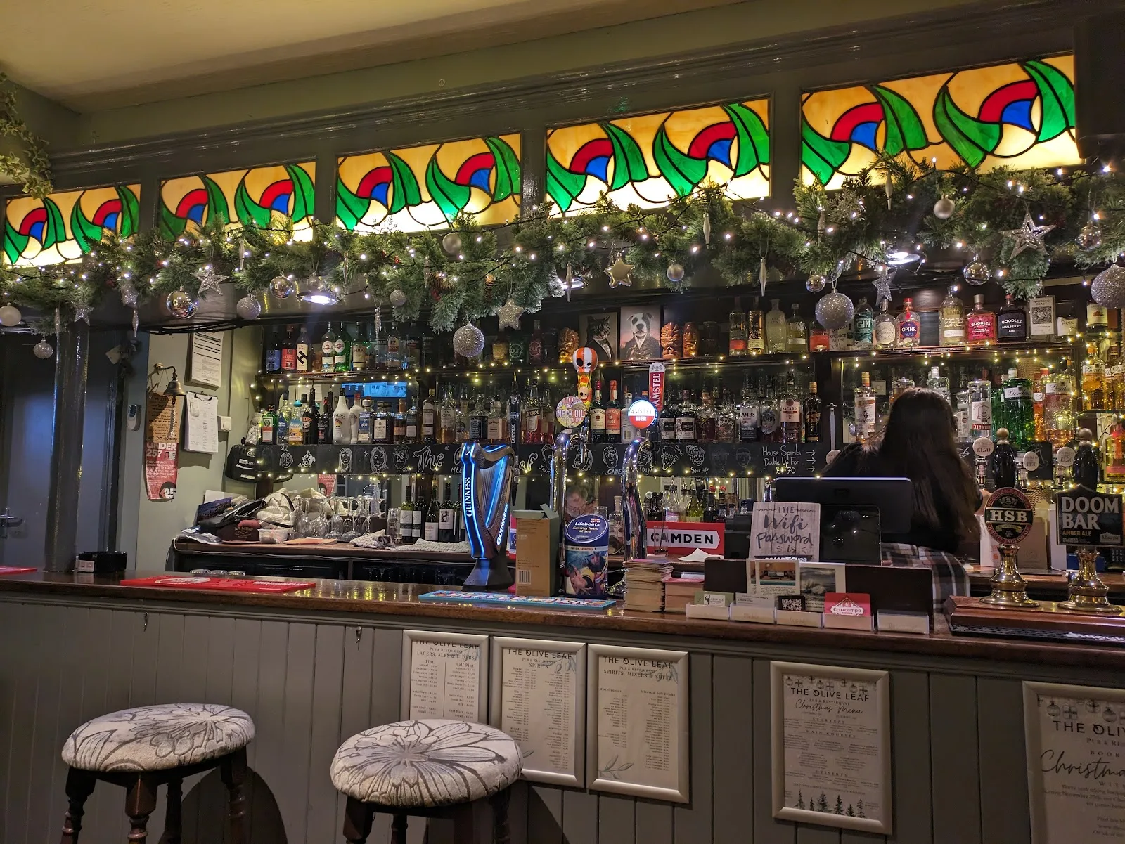 The bar at The Olive Leaf with stained glass panels, a full spirits shelf and hand pumps, dressed for Christmas